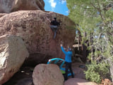 A climber near the top of a sandstone boulder, a spotter and crash pad below